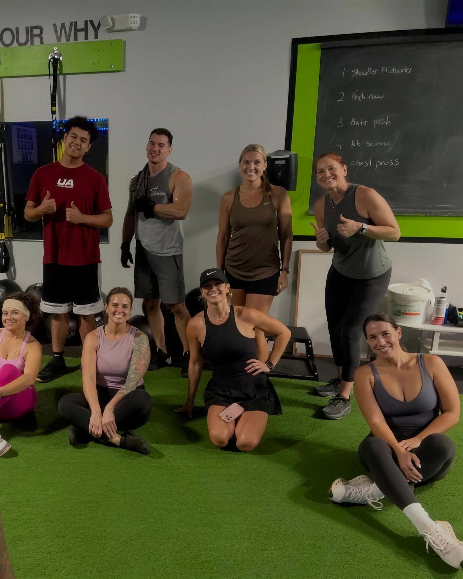 A Sunday class sitting together on the turf after a workout, smiling at the camera